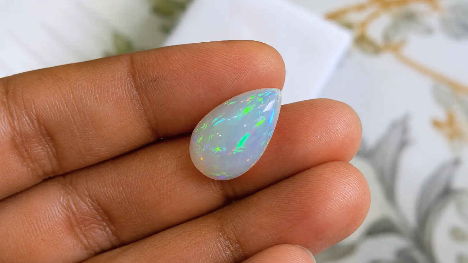 A pear-shaped natural opal gemstone is held between fingertips, revealing its colorful play-of-color with vivid green and blue flashes Opal Wedding Rings