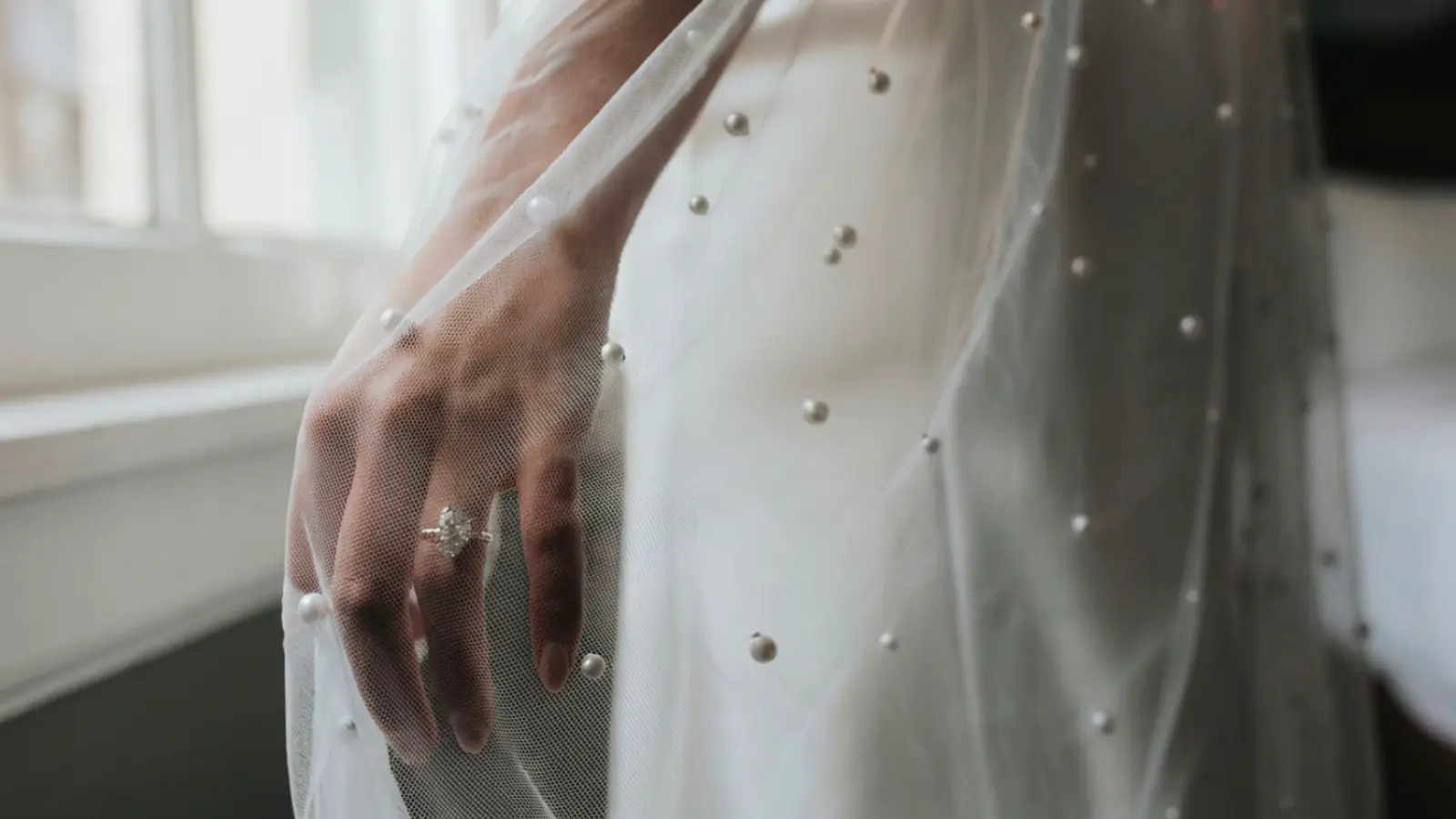Bride's hand wearing an engagement ring beneath a pearl wedding veil with scattered pearl embellishments