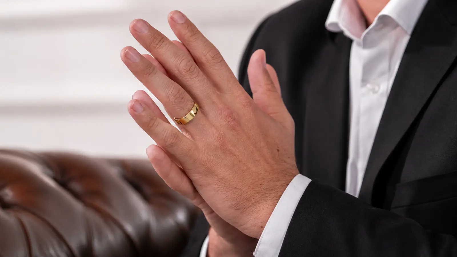 Close-up of a groom wearing a polished yellow gold men's wedding band with a black tuxedo and white dress shirt