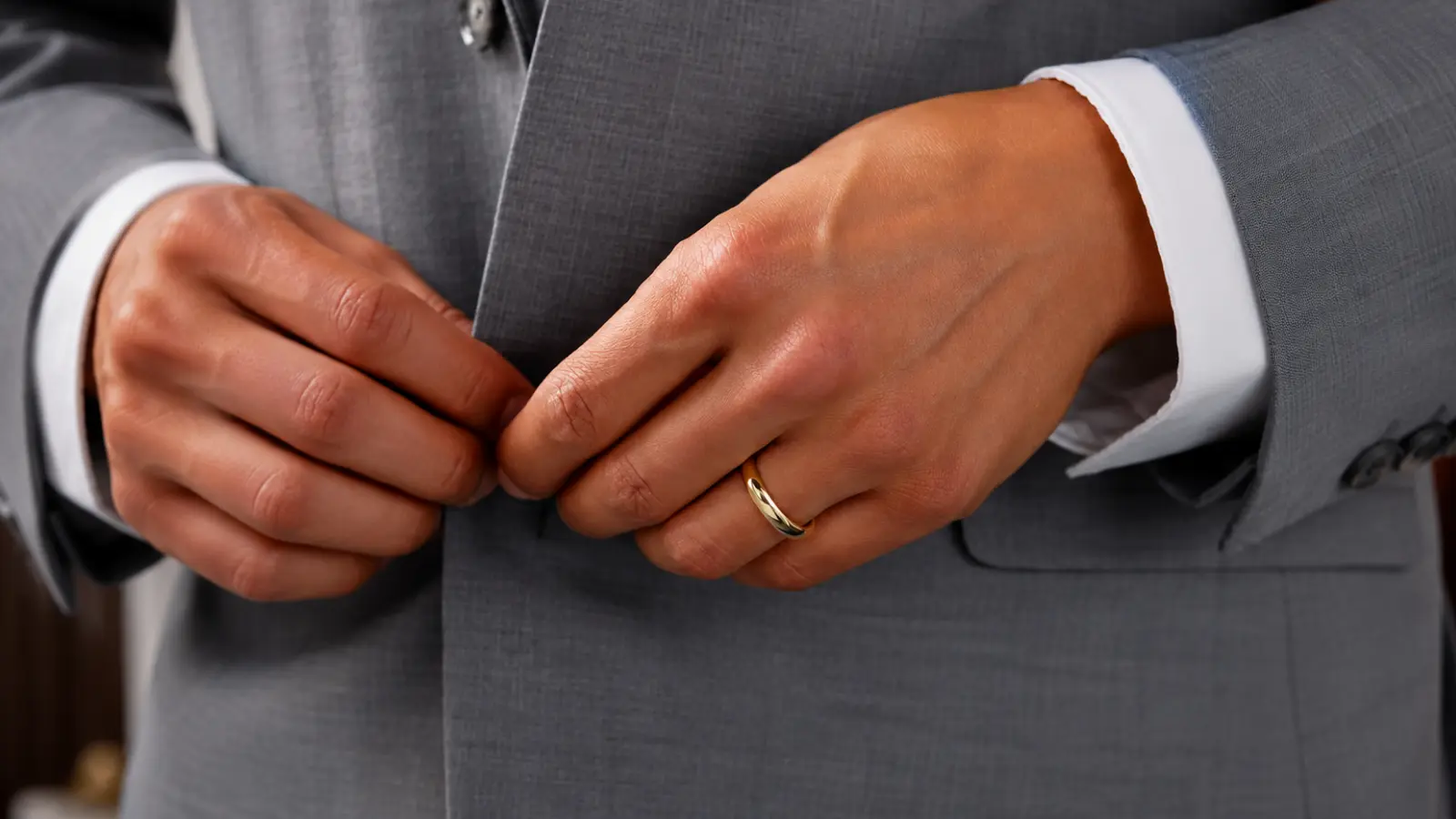 Groom fastening a gray suit jacket while wearing a polished yellow gold men's wedding band, illustrating a timeless and elegant wedding ring that complements formal attire
