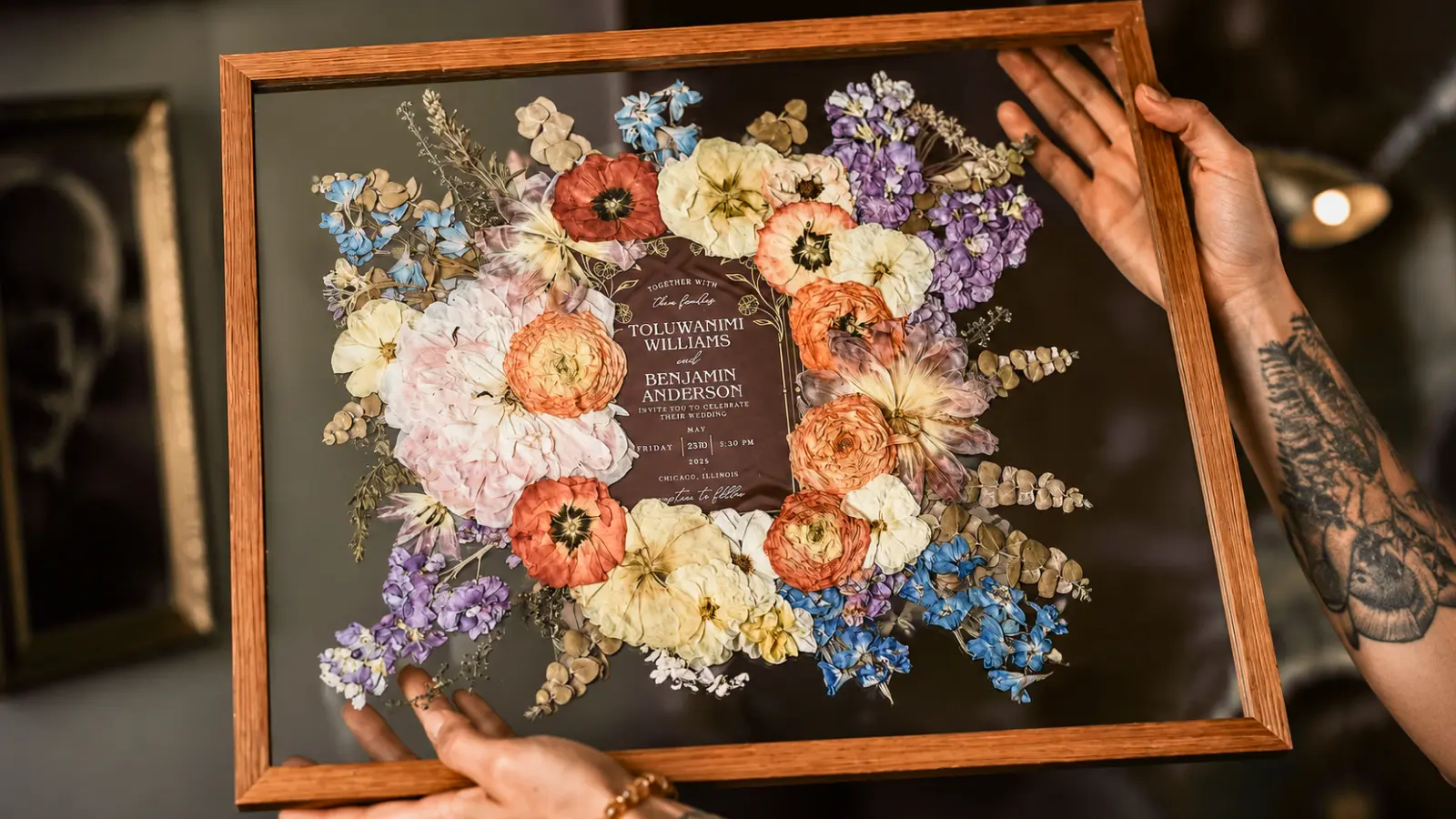 Hands holding a wooden frame with colorful pressed wedding flowers arranged around a preserved invitation