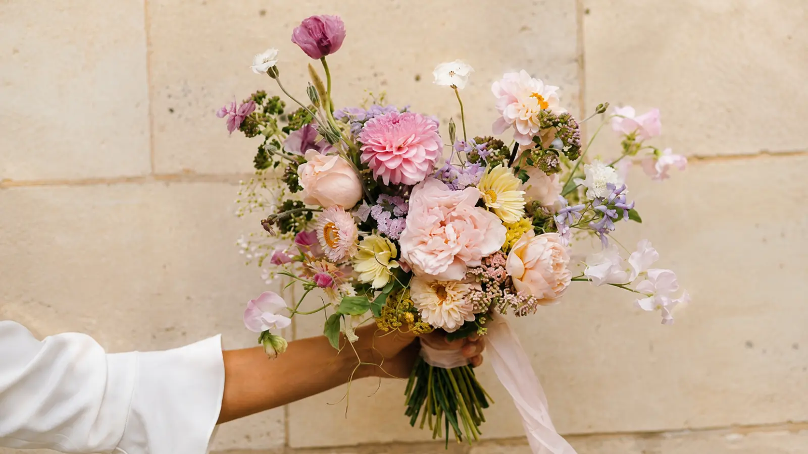 Hand holding a romantic spring garden wedding bouquet with blush roses, dahlias and pastel seasonal flowers
