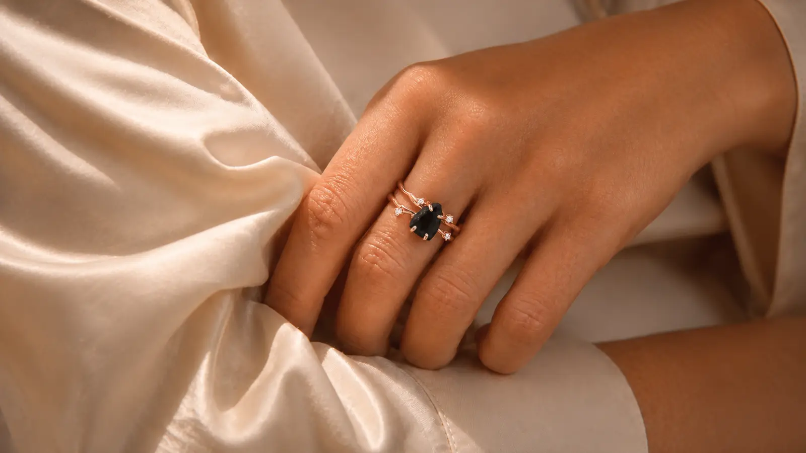 A hand resting on satin fabric while wearing a rose gold gothic black engagement ring with an oval black gemstone