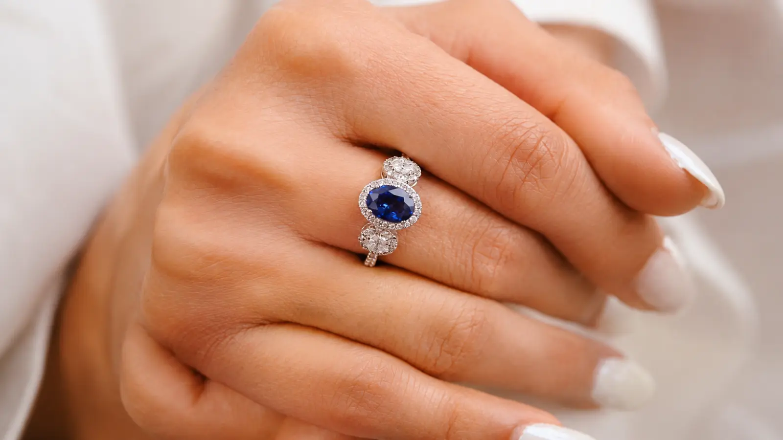 A close-up of a sapphire three-stone halo engagement ring featuring a blue center stone framed by diamonds on a white metal band