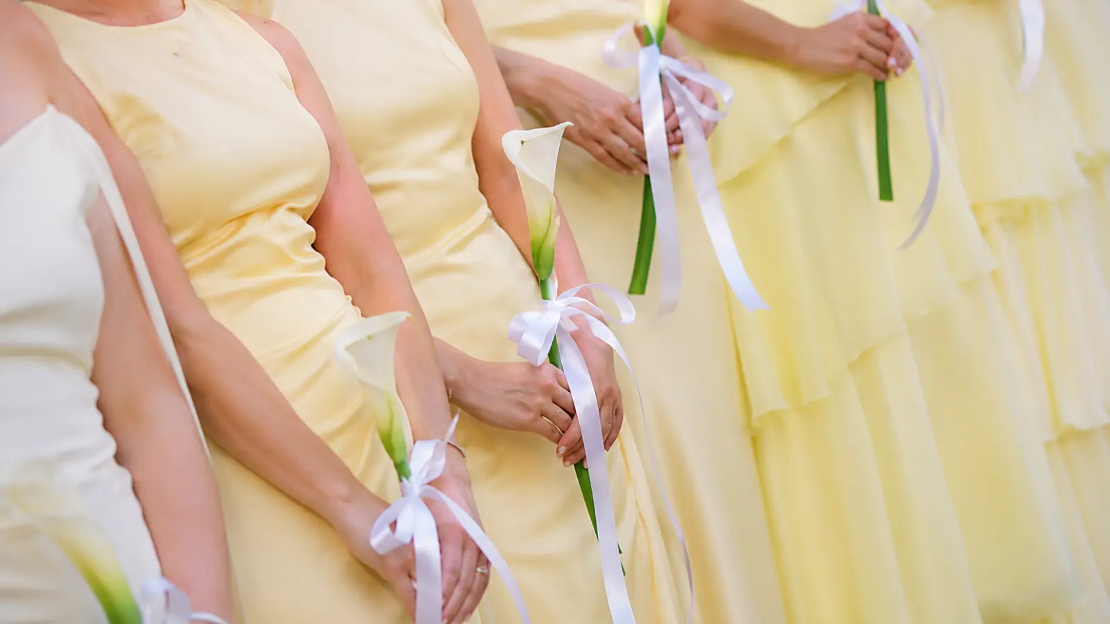 Bridesmaids in yellow dresses holding single white calla lily bouquets with ribbons