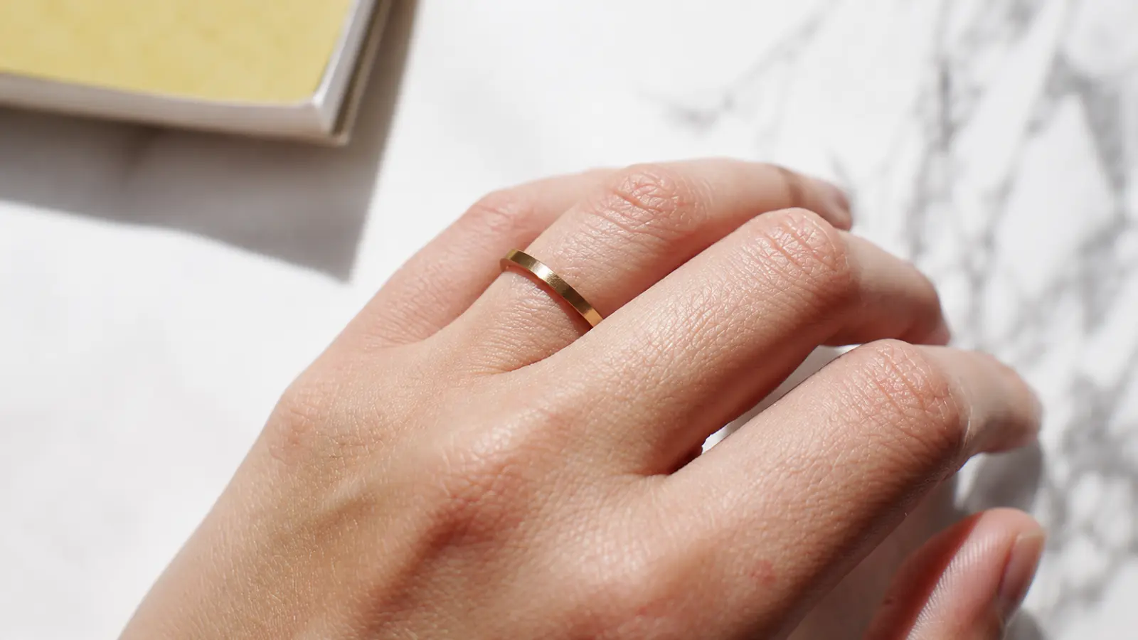 A hand wearing a slim classic yellow gold wedding band with a clean minimalist profile against a bright marble background