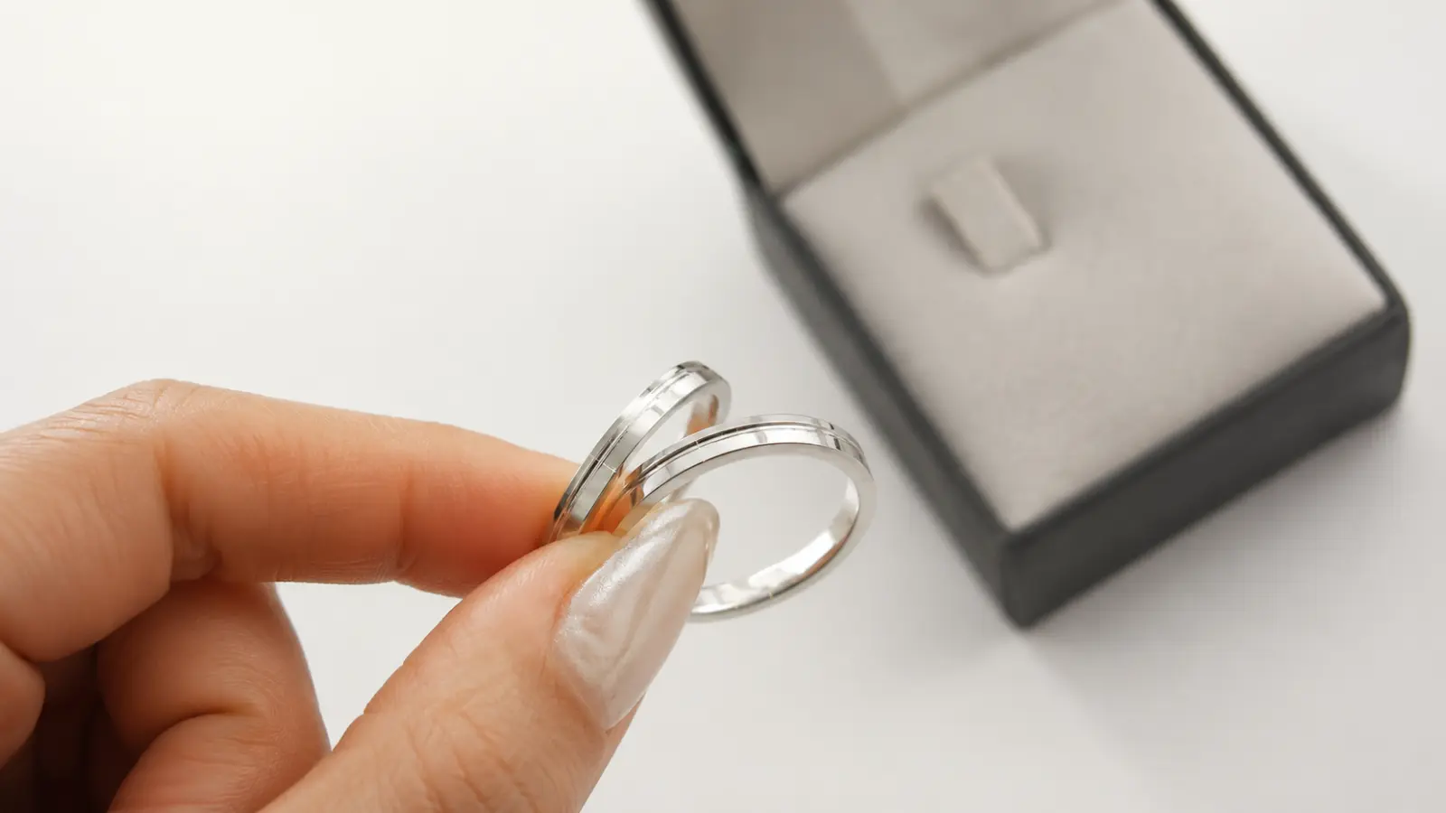 A hand holds two slim silver wedding rings near an open jewelry box, emphasizing their polished surfaces, elegant proportions and timeless wedding band design
