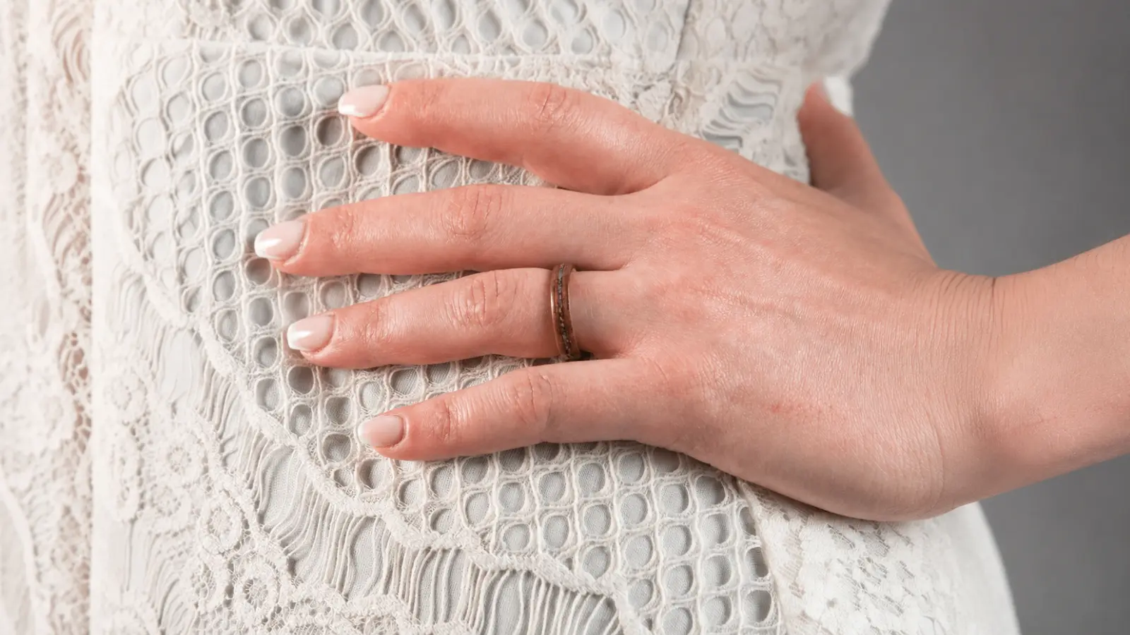 A bride's hand rests on a lace wedding dress while wearing a slim wood wedding ring, emphasizing the organic grain and refined minimalist design