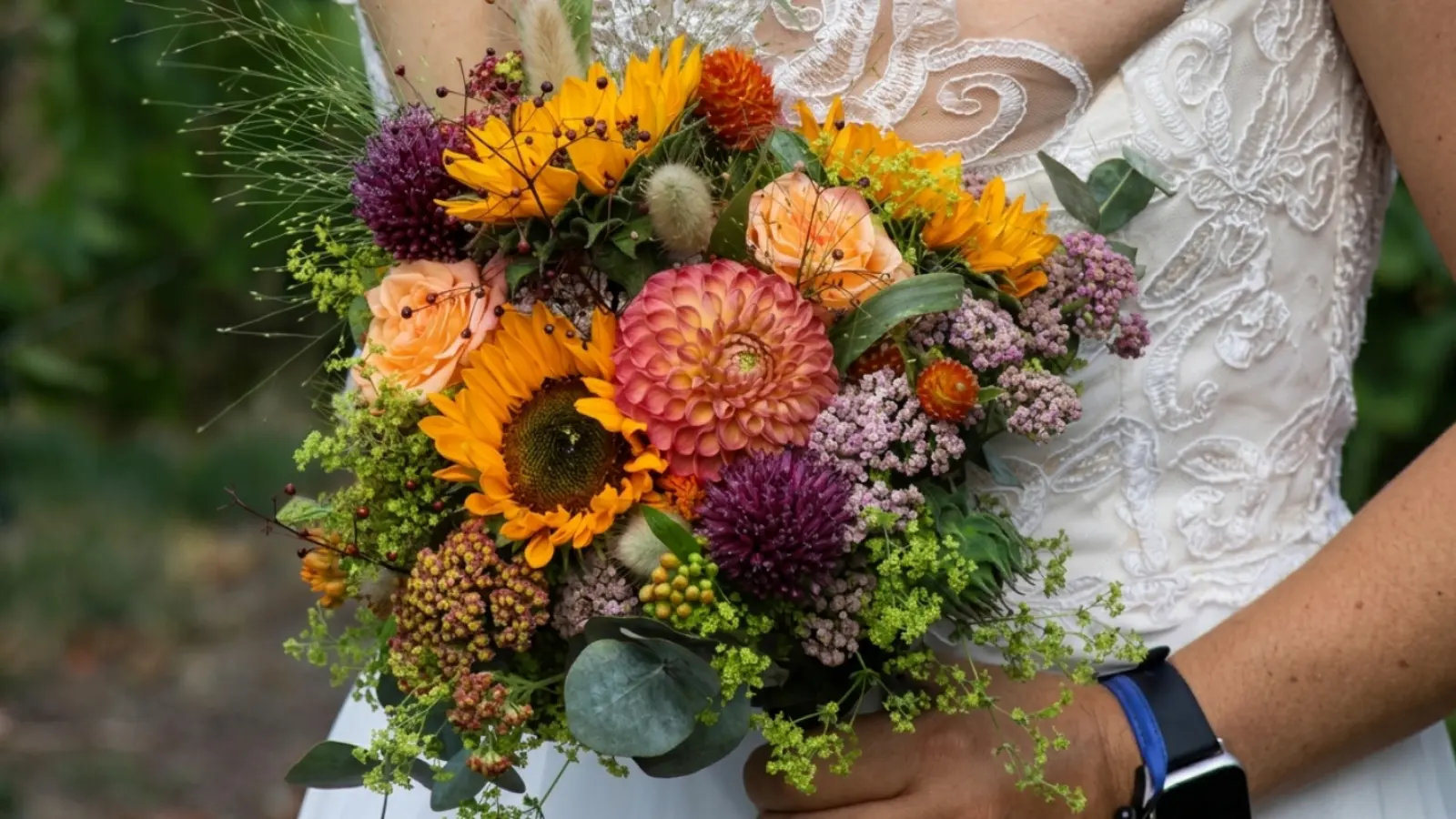 Bride holding a fall sunflower wedding bouquet with dahlias, roses, allium, and textured greenery