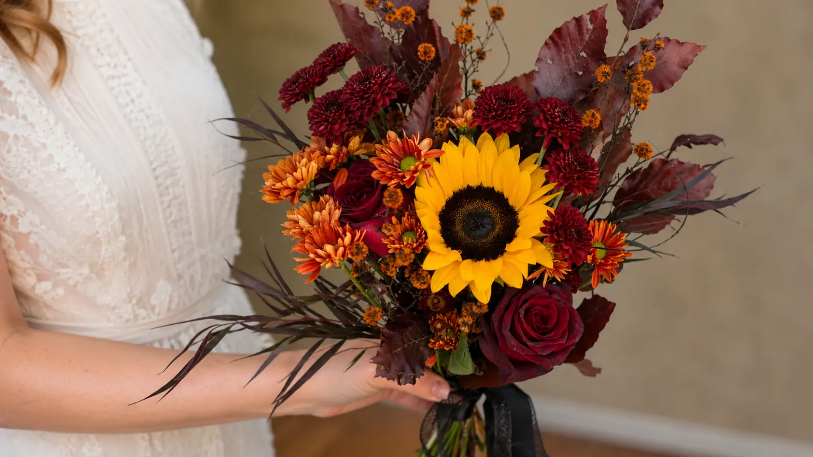 Bride holding a fall wedding bouquet with a sunflower, burgundy roses and rich autumn flowers