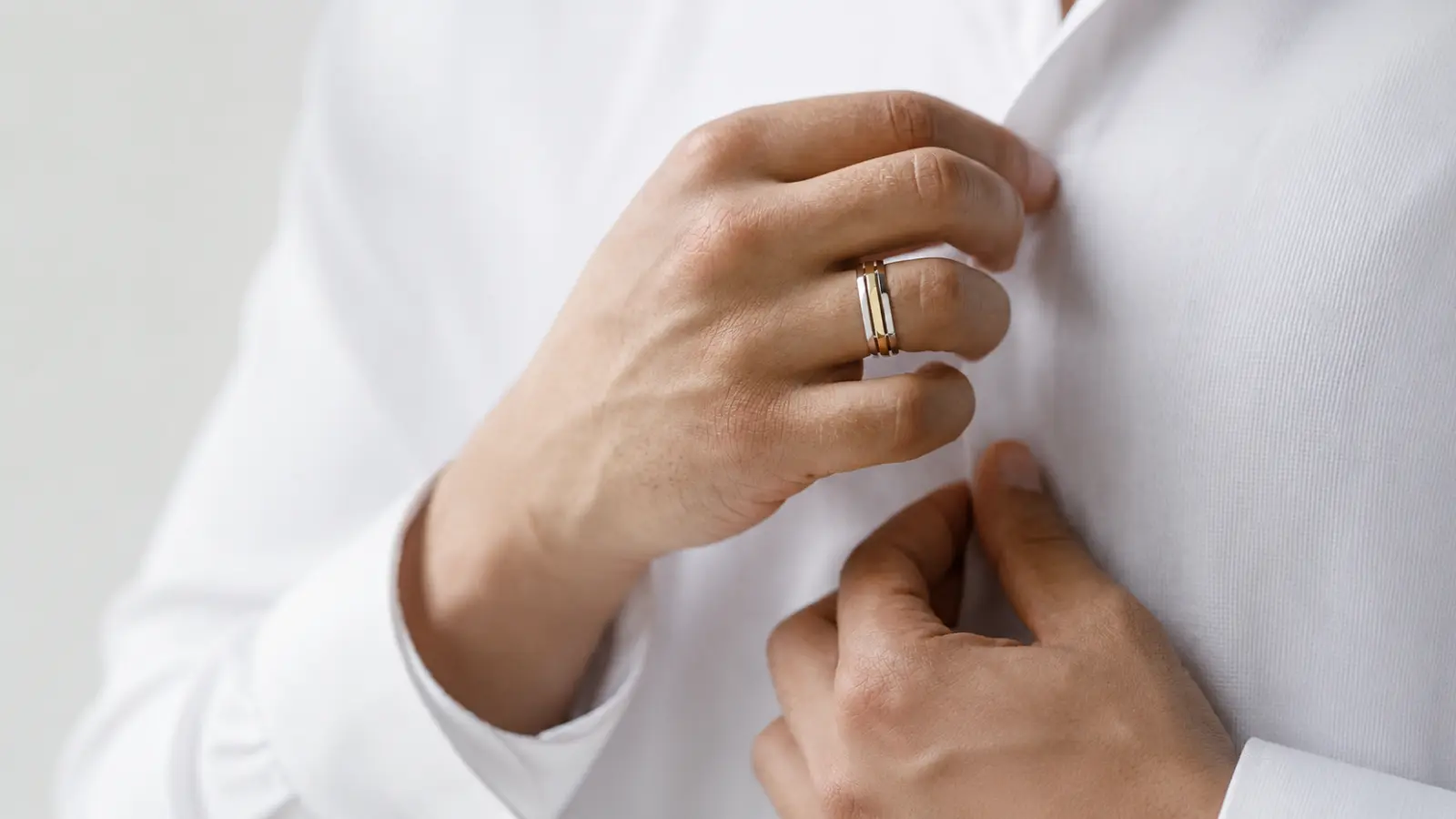 Close-up of a man fastening a white dress shirt while wearing a modern two-tone men's wedding band, highlighting the clean lines and contemporary mixed-metal design