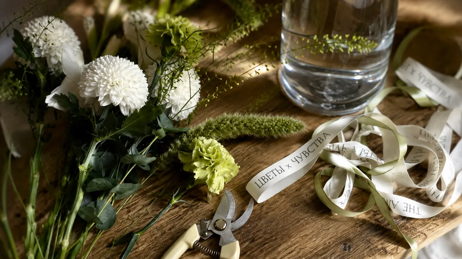 Fresh flowers, floral ribbon, pruning shears, and a vase of water arranged on a wooden table for making a wedding bouquet