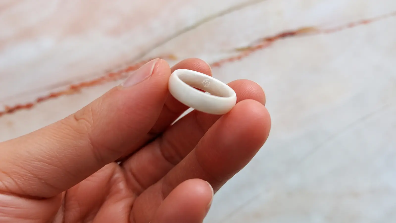 A close-up of a white silicone wedding ring held between two fingers, showcasing its smooth texture, flexible material and simple modern appearance