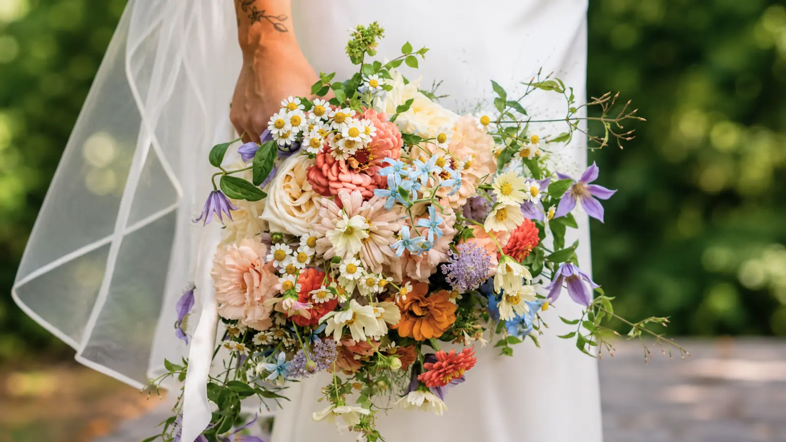 Bride holding a mixed wildflower wedding bouquet with daisies, roses, clematis, and pastel seasonal flowers