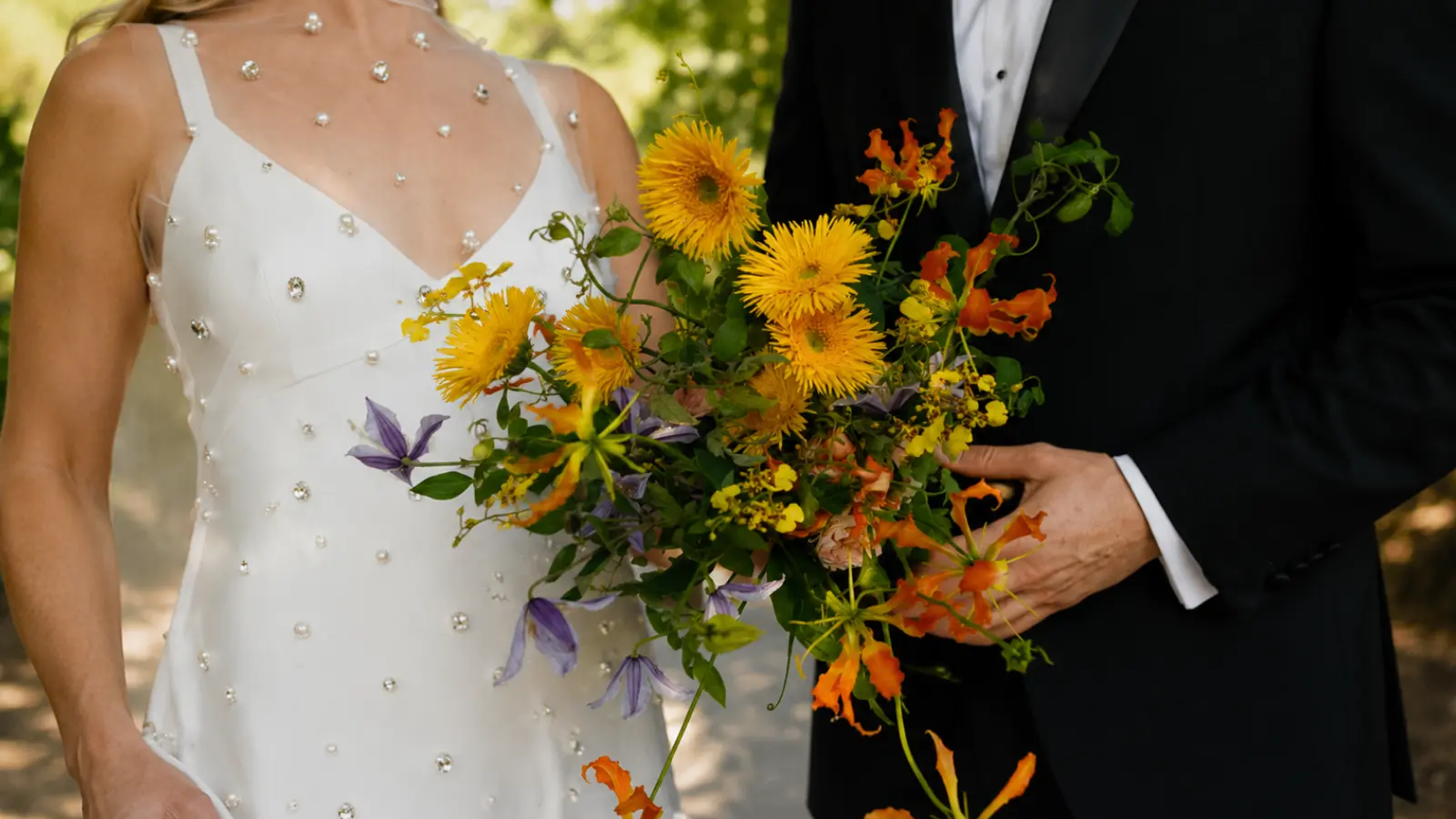 Bride and groom holding a yellow wildflower wedding bouquet with orange lilies, purple flowers, greenery, and bright garden blooms
