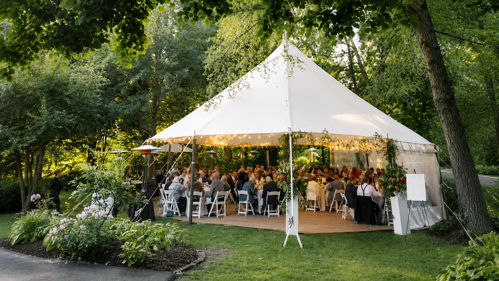 Backyard wedding reception under a large white tent with seated guests, dining tables and surrounding trees and landscaping