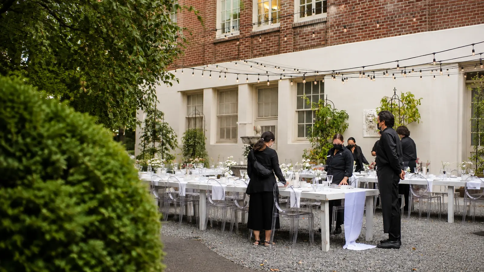 Event staff setting up tables, chairs and place settings for a backyard wedding reception beside a residential building