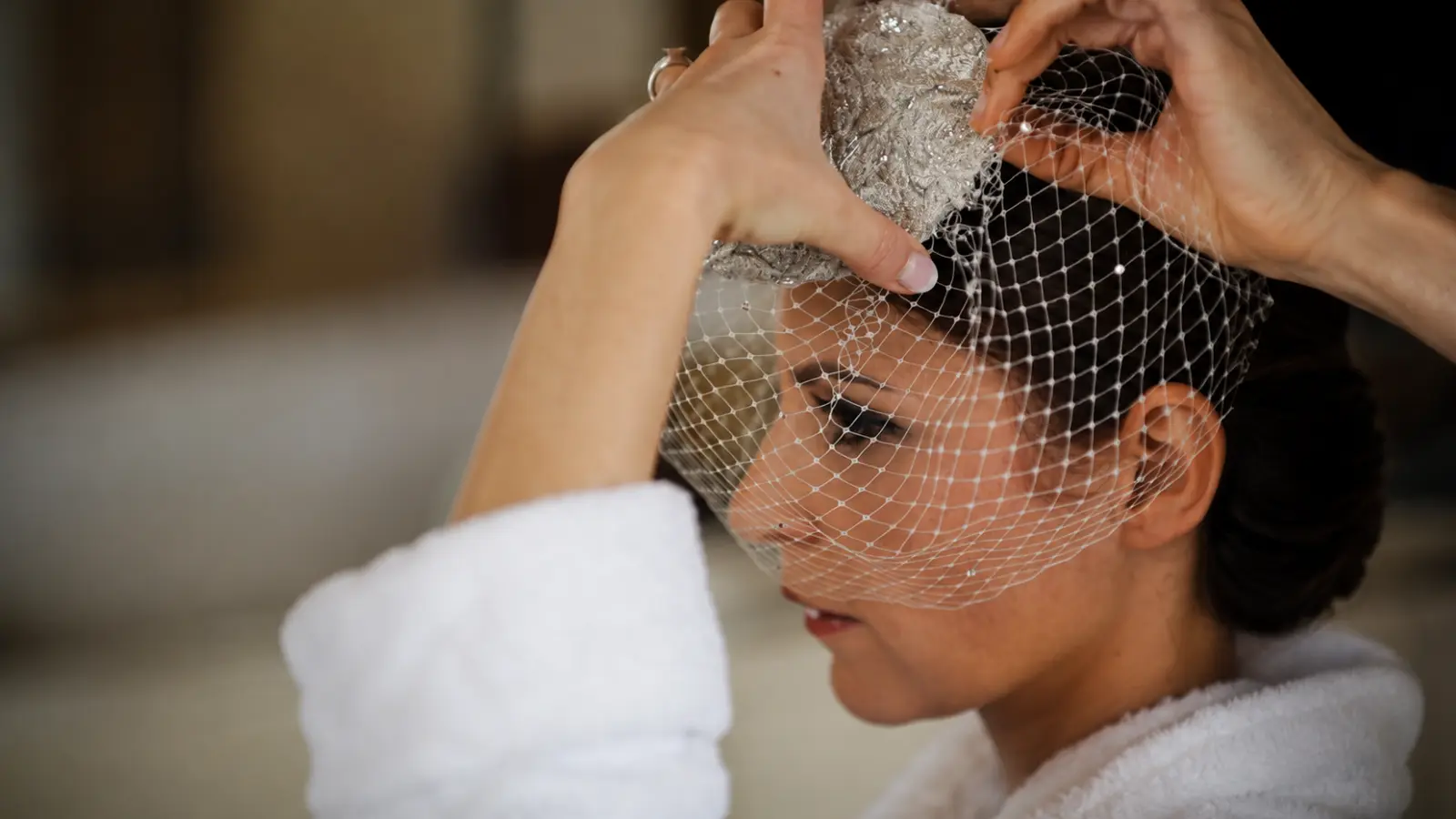 Bride adjusting a white birdcage wedding veil over her face while positioning the netting around a decorative bridal headpiece
