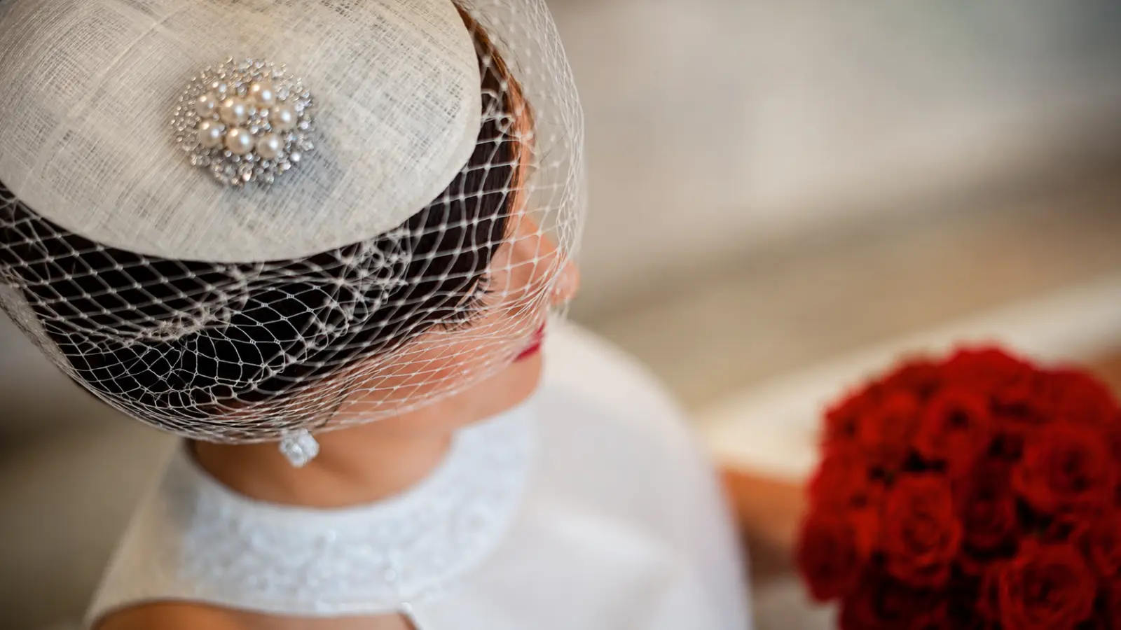 Bride wearing a white birdcage wedding veil with an ivory fascinator and pearl embellishment while holding a bouquet of red roses