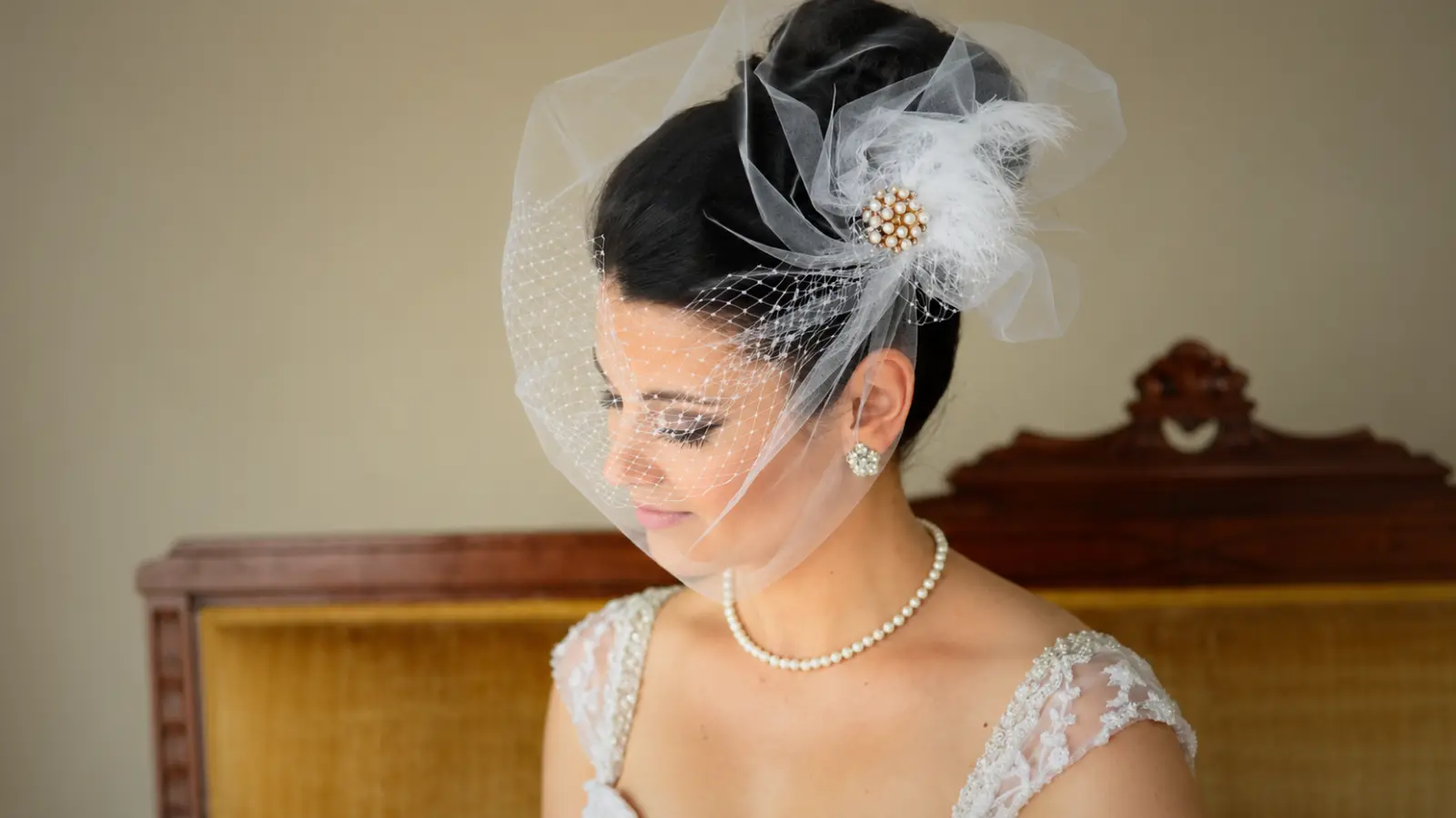 Bride wearing a white birdcage wedding veil over her face with a pearl and feather fascinator attached beside her bridal updo