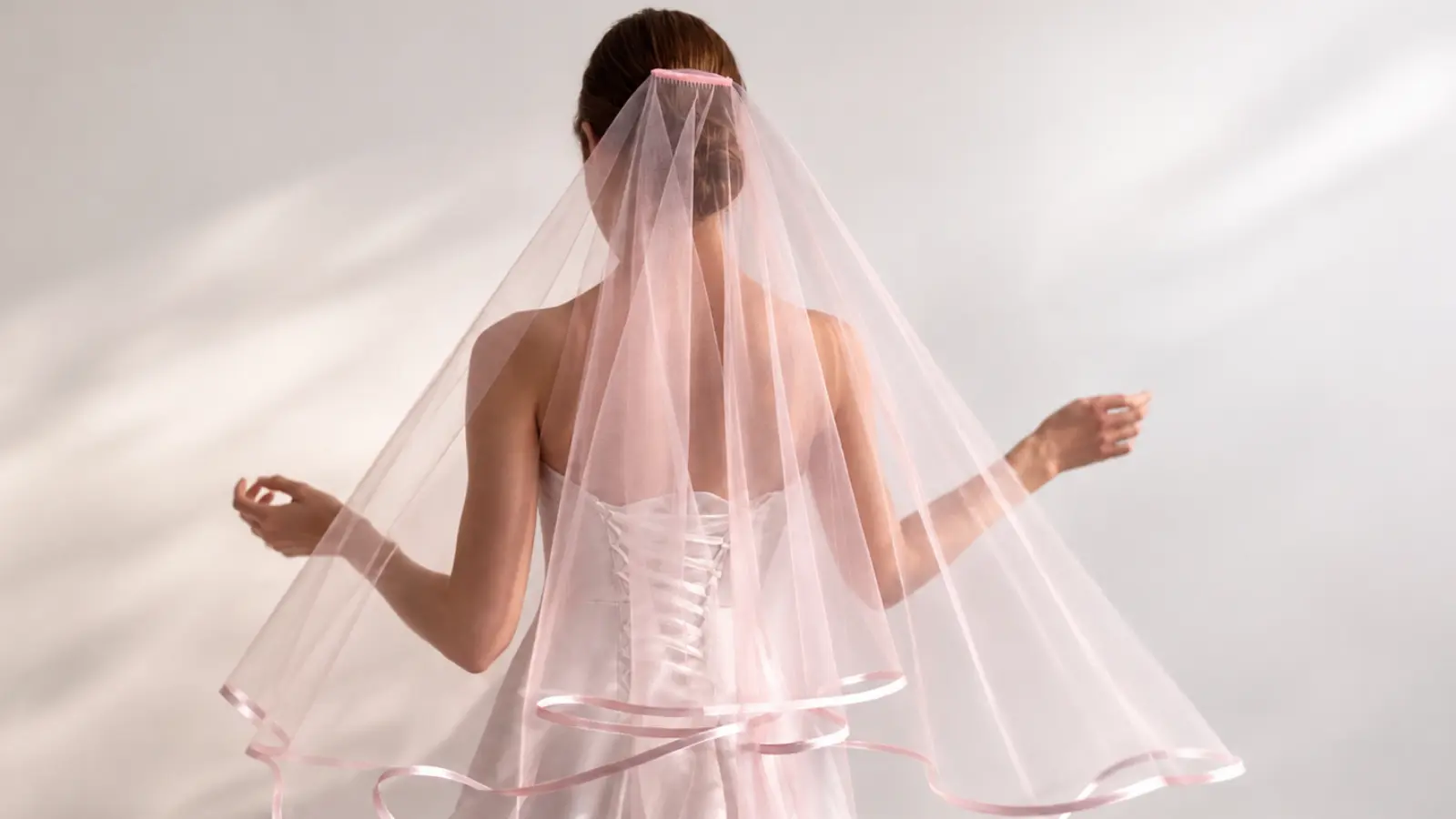 Bride wearing a blush pink colored wedding veil with sheer tulle and delicate satin ribbon edging over a white wedding dress