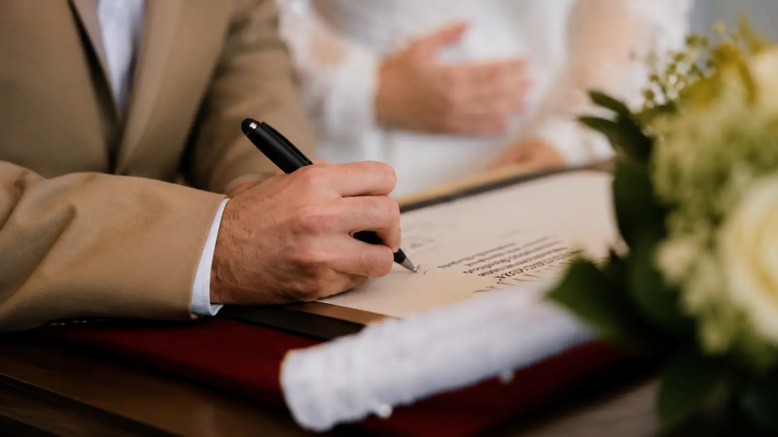 Groom signing official marriage paperwork beside the bride with wedding flowers nearby during the marriage documentation process Certified Copy of a Marriage Certificate: Meaning and Uses