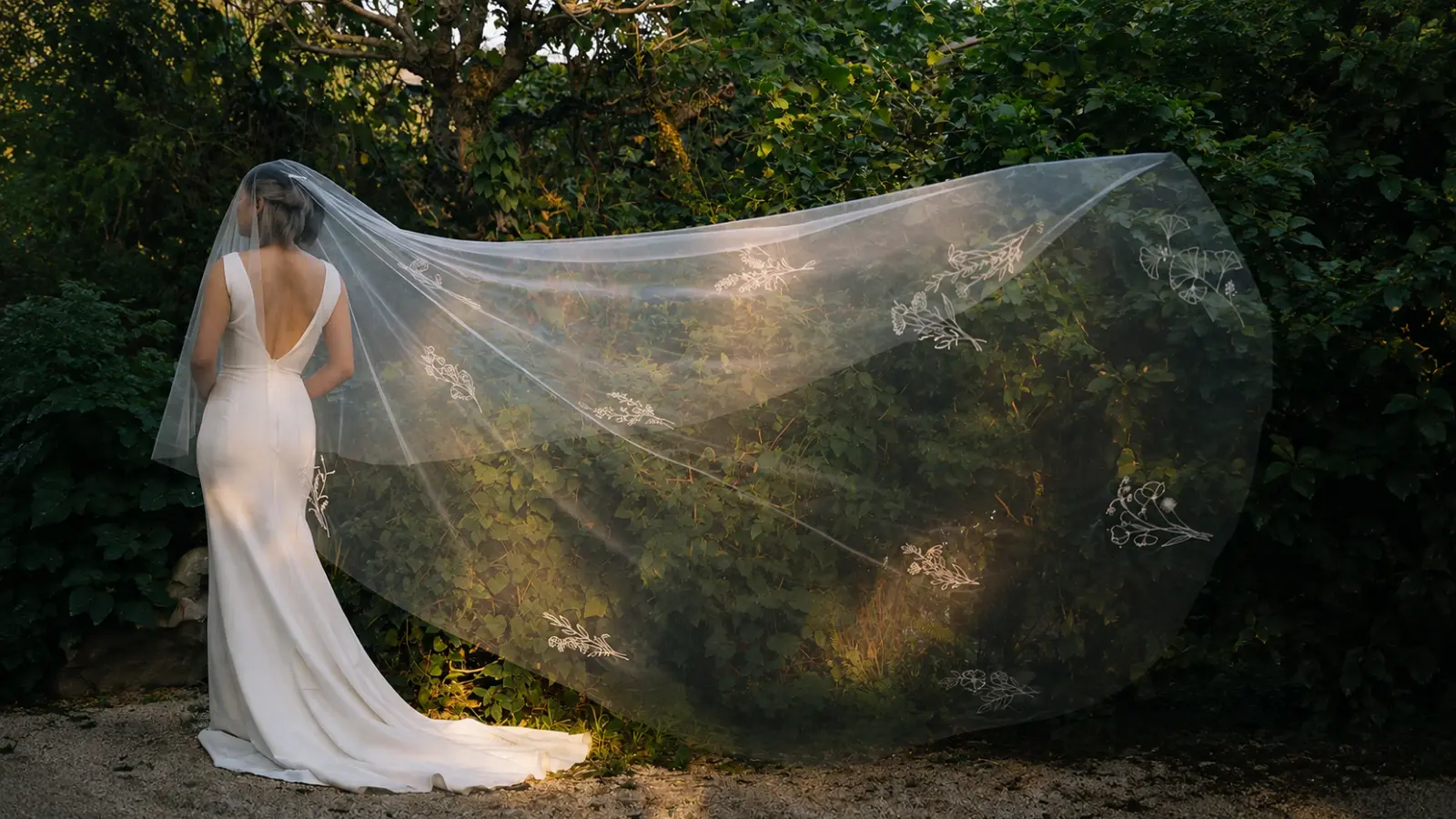 Bride wearing a sheer chapel length wedding veil flowing dramatically behind her during an outdoor wedding portrait in a grassy landscape