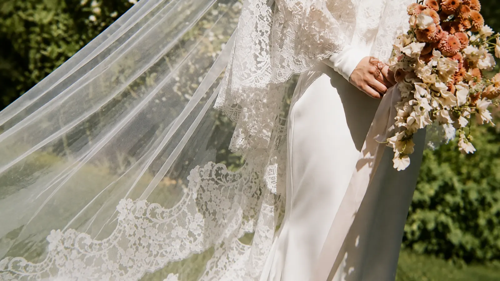 Bride wearing a long chapel length wedding veil with ornate lace trim flowing behind her while carrying a bouquet of white roses