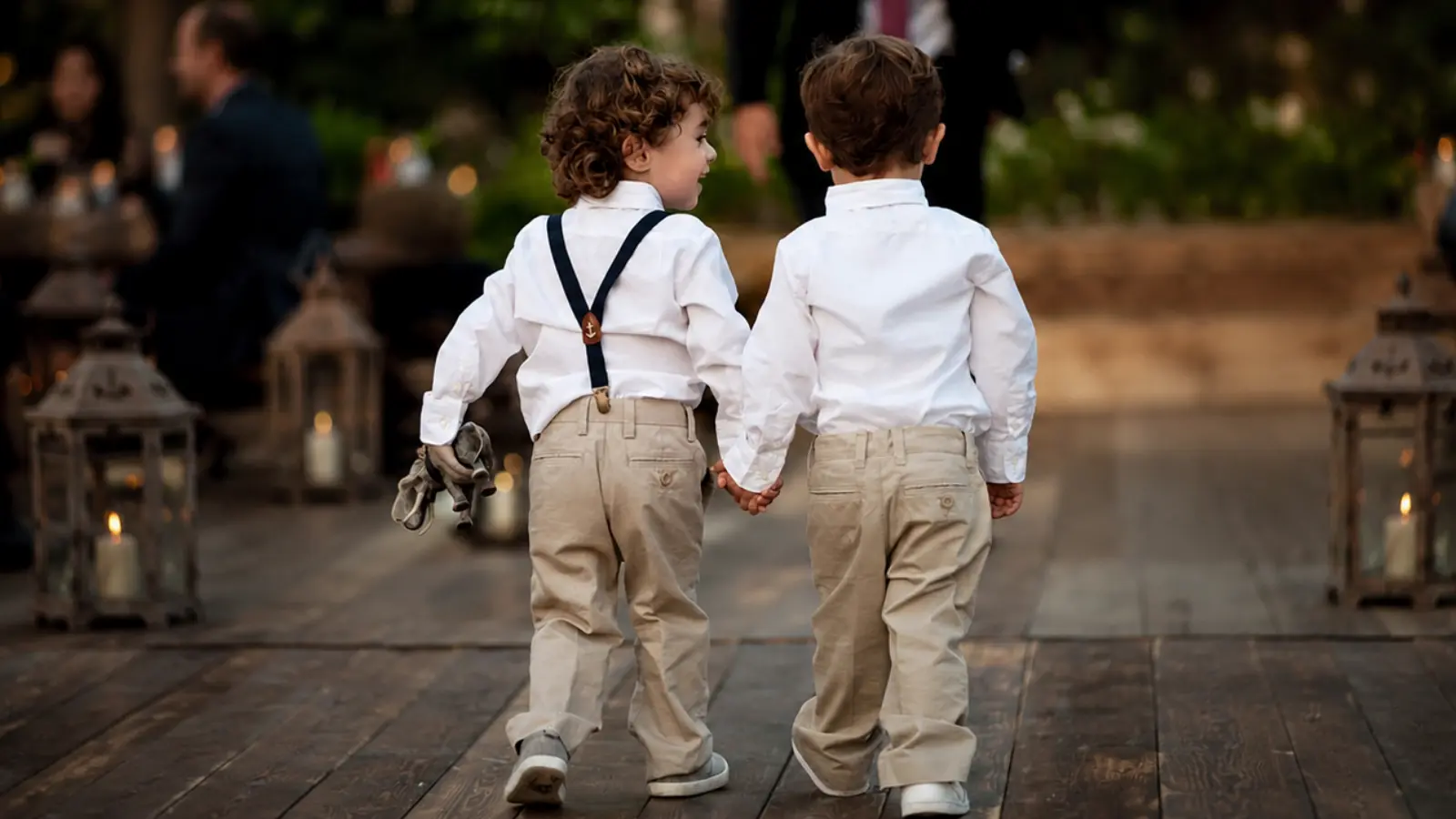 Two young ring bearers walking hand in hand in coordinated white shirts and beige pants during an outdoor wedding celebration