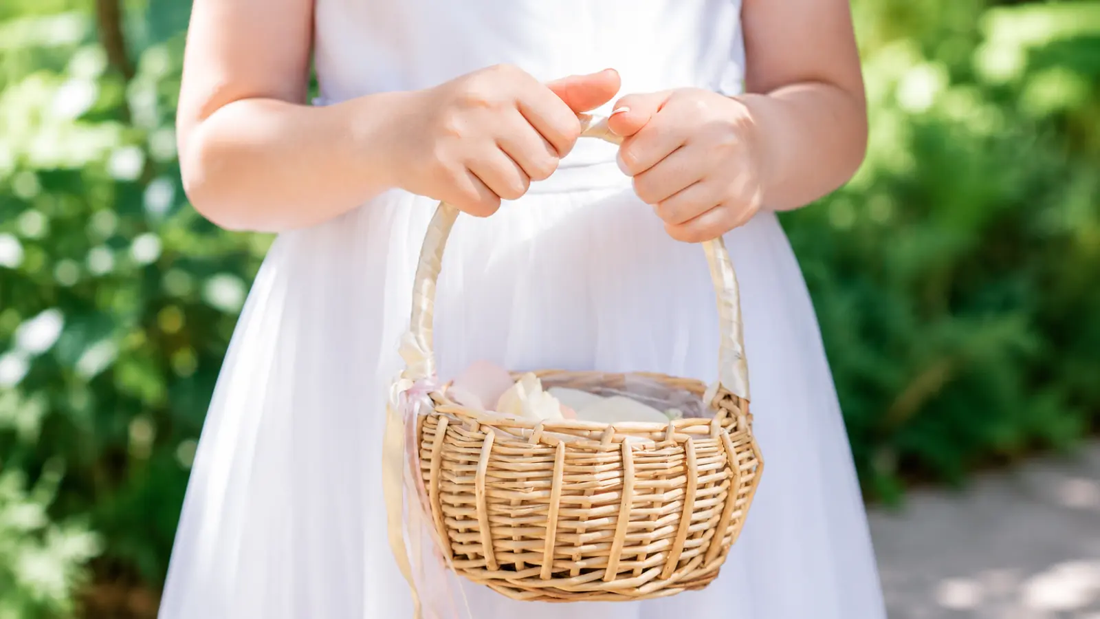 Flower girl basket made of natural wicker with a wrapped handle and soft flower petals carried by a girl in a white wedding dress