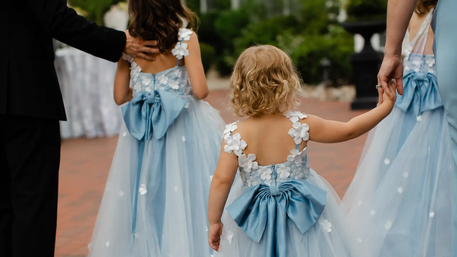 Flower girl dress colors shown in light blue dresses with white floral appliqués, tulle skirts, and large blue bows at a wedding