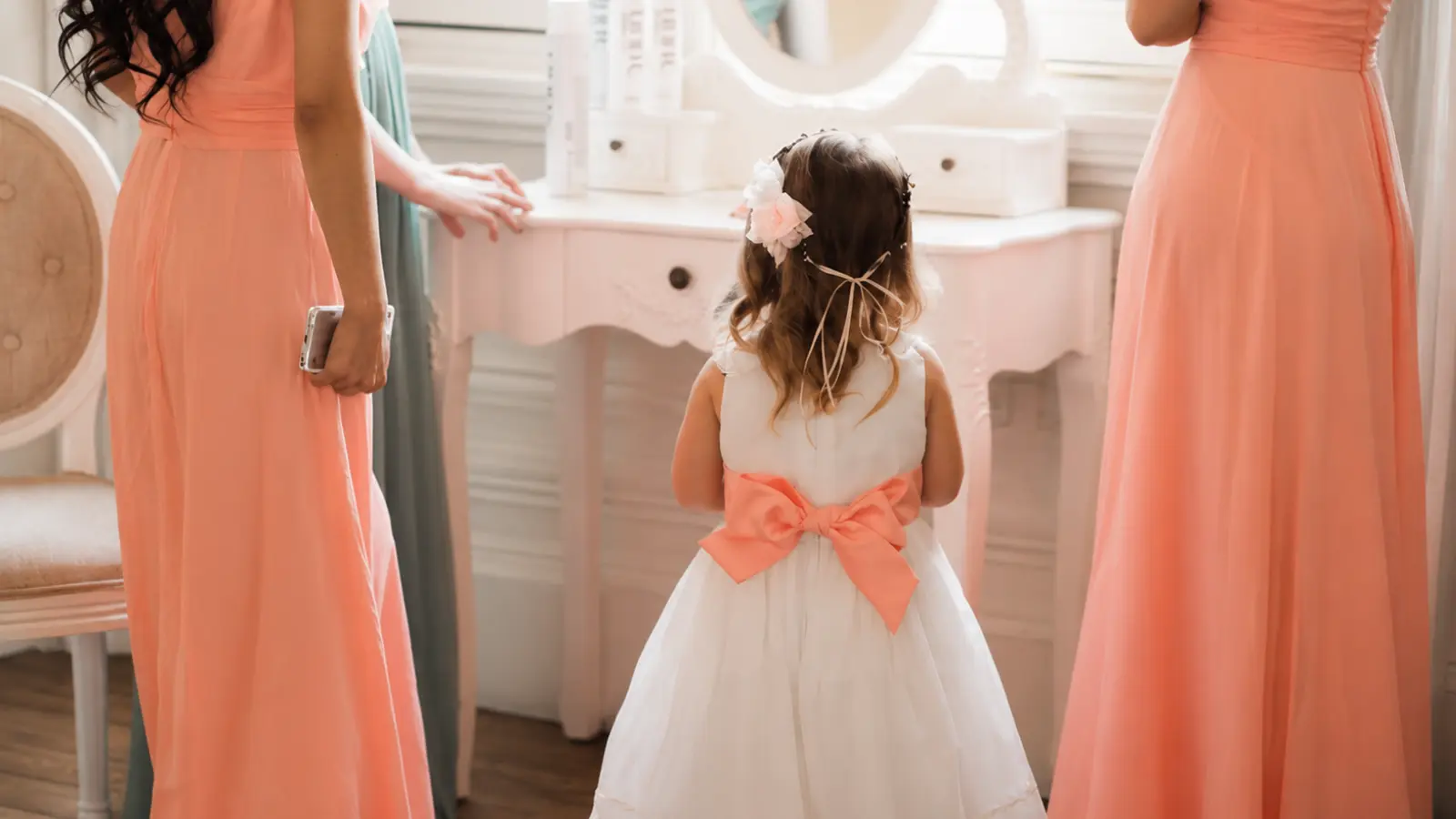 Flower girl dress colors shown with a white dress and coral bow coordinated with coral bridesmaid dresses in a bright wedding getting-ready room