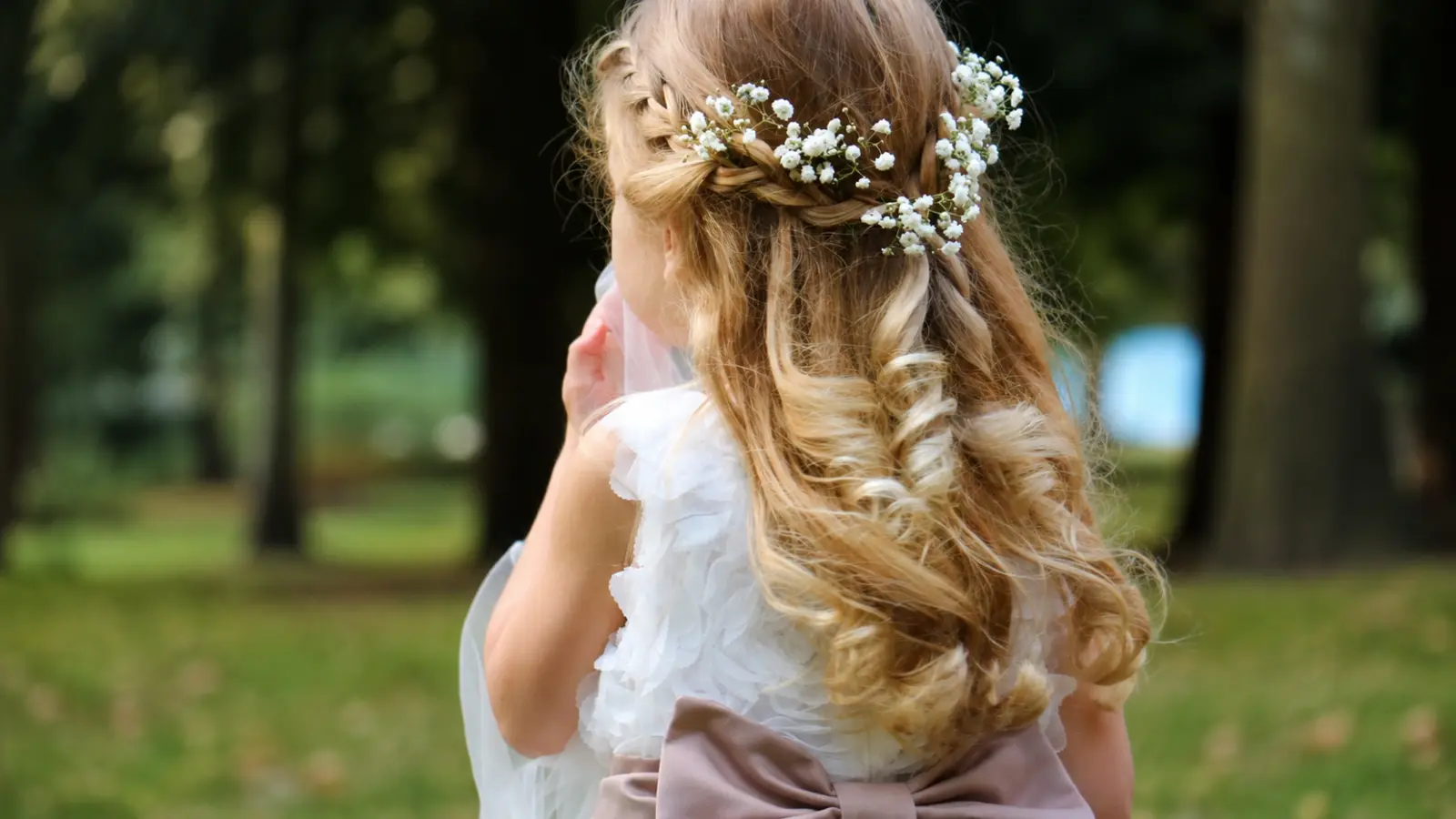 Flower girl hairstyle with long blonde curls, a braided crown, and small white baby’s breath flowers arranged throughout the hair