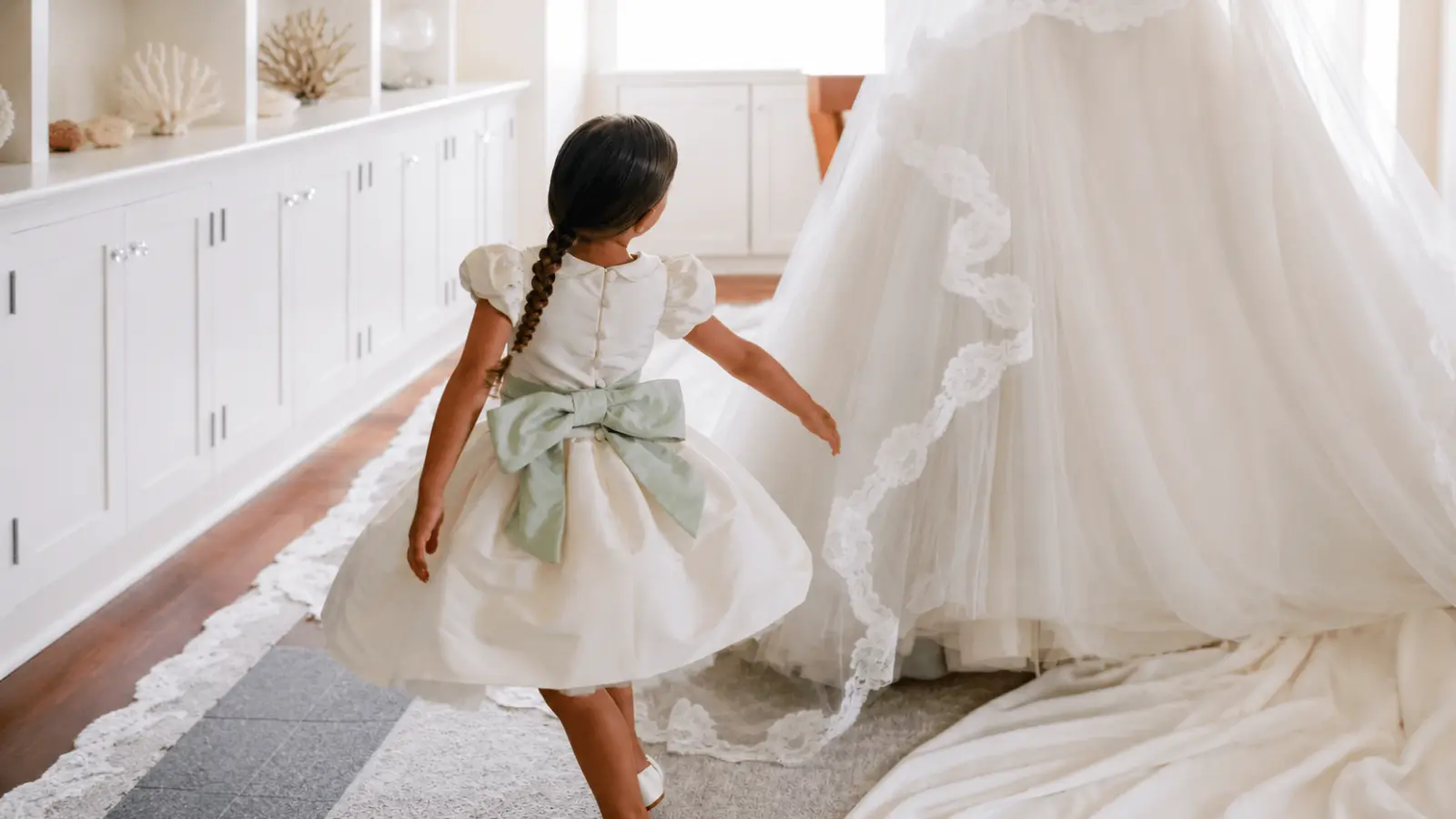Flower girl in a white dress with a pale green bow walks beside the bride and her gown before the wedding ceremony