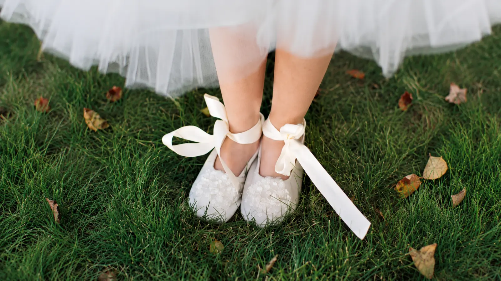 Flower girl shoes with white floral details and long ribbon ankle ties worn beneath a tulle dress on grass at an outdoor wedding