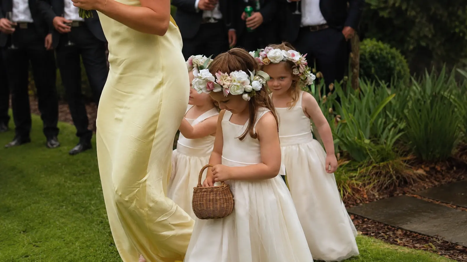 Flower girls in white dresses and floral crowns walk together with a small basket during an outdoor wedding ceremony processional