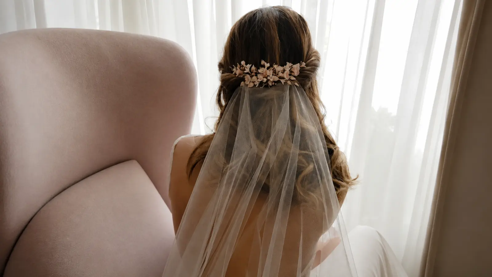 Curled half up half down wedding hair with a sheer veil positioned beneath a delicate floral hairpiece at the back of the hairstyle