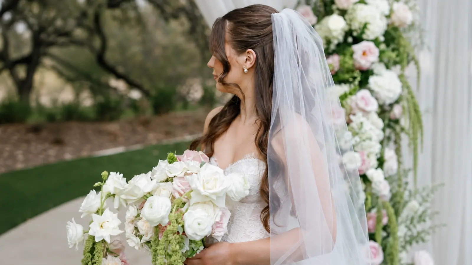 Bride with long half up half down wedding hair wears a sheer veil while holding a white and blush floral wedding bouquet