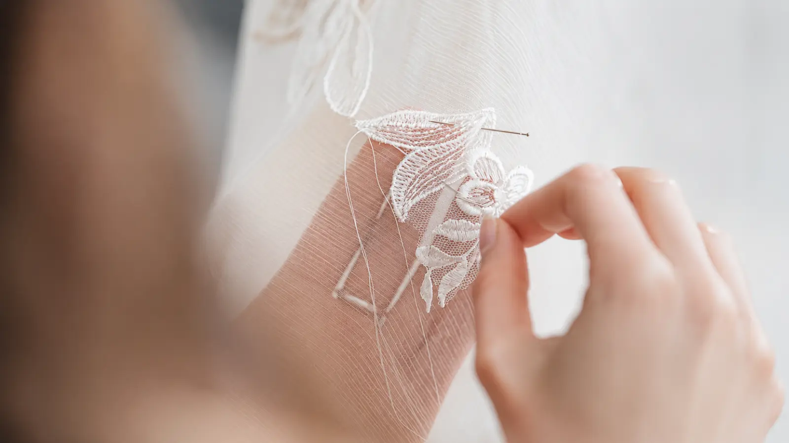 Artisan hand-sewing floral lace appliqué onto sheer wedding veil fabric, demonstrating detailed craftsmanship that can increase the cost of a bridal veil Wedding Veil Cost
