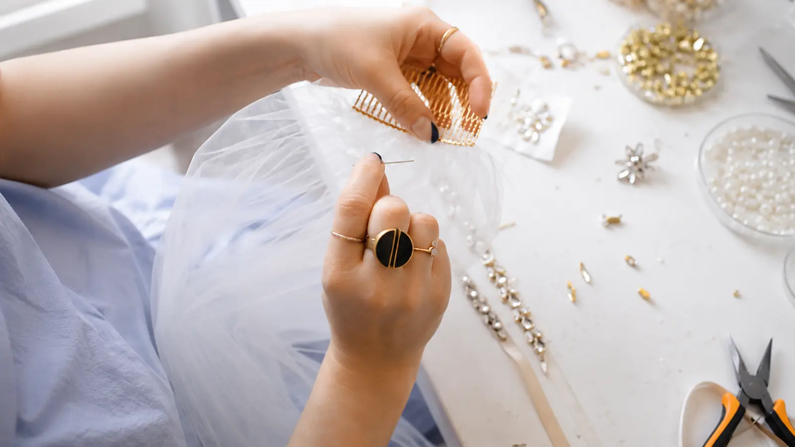 Hands sewing a gold comb onto gathered white tulle during the process of making a DIY wedding veil at home How to Make a Wedding Veil
