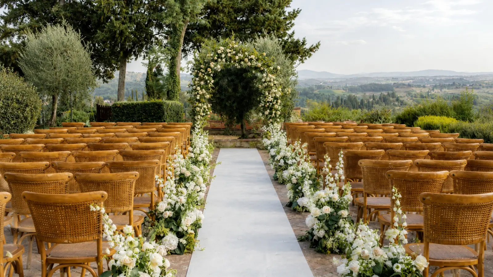 Outdoor wedding ceremony setup with wooden guest chairs, white floral aisle arrangements and a flower-covered ceremony arch overlooking the countryside