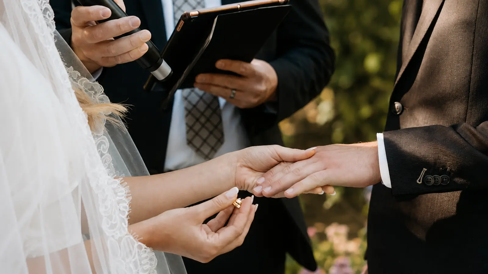 Bride placing a wedding ring on the groom’s hand while an officiant leads the ring exchange during an outdoor wedding ceremony