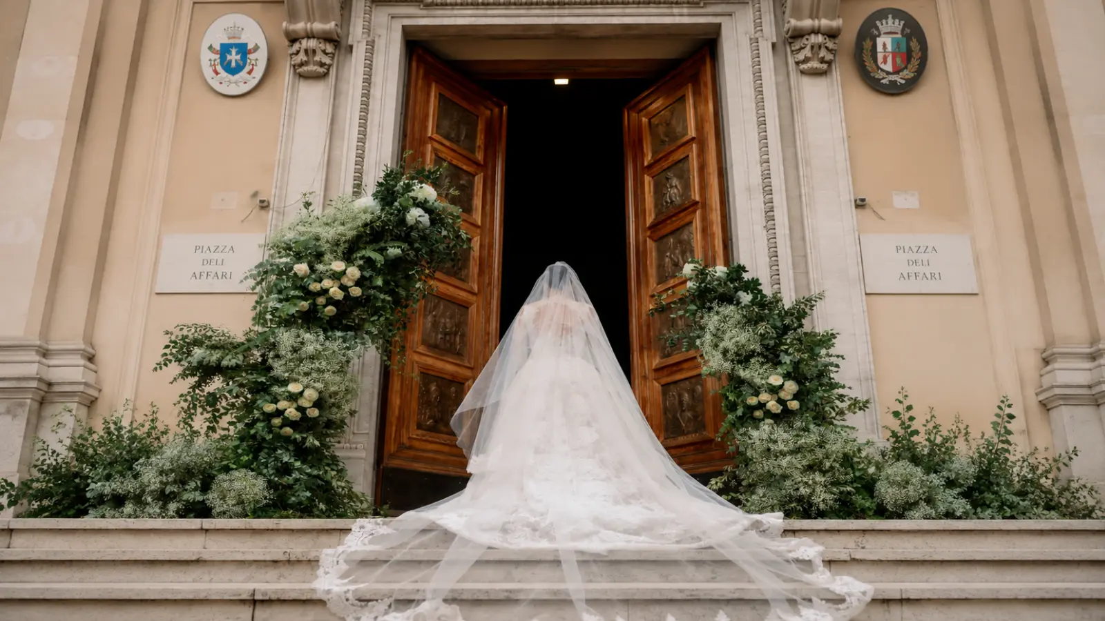 Bride wearing a long veil entering a formal wedding ceremony venue with large wooden doors, white floral arrangements and greenery How to Plan a Wedding Ceremony: Step-by-Step Planning Guide