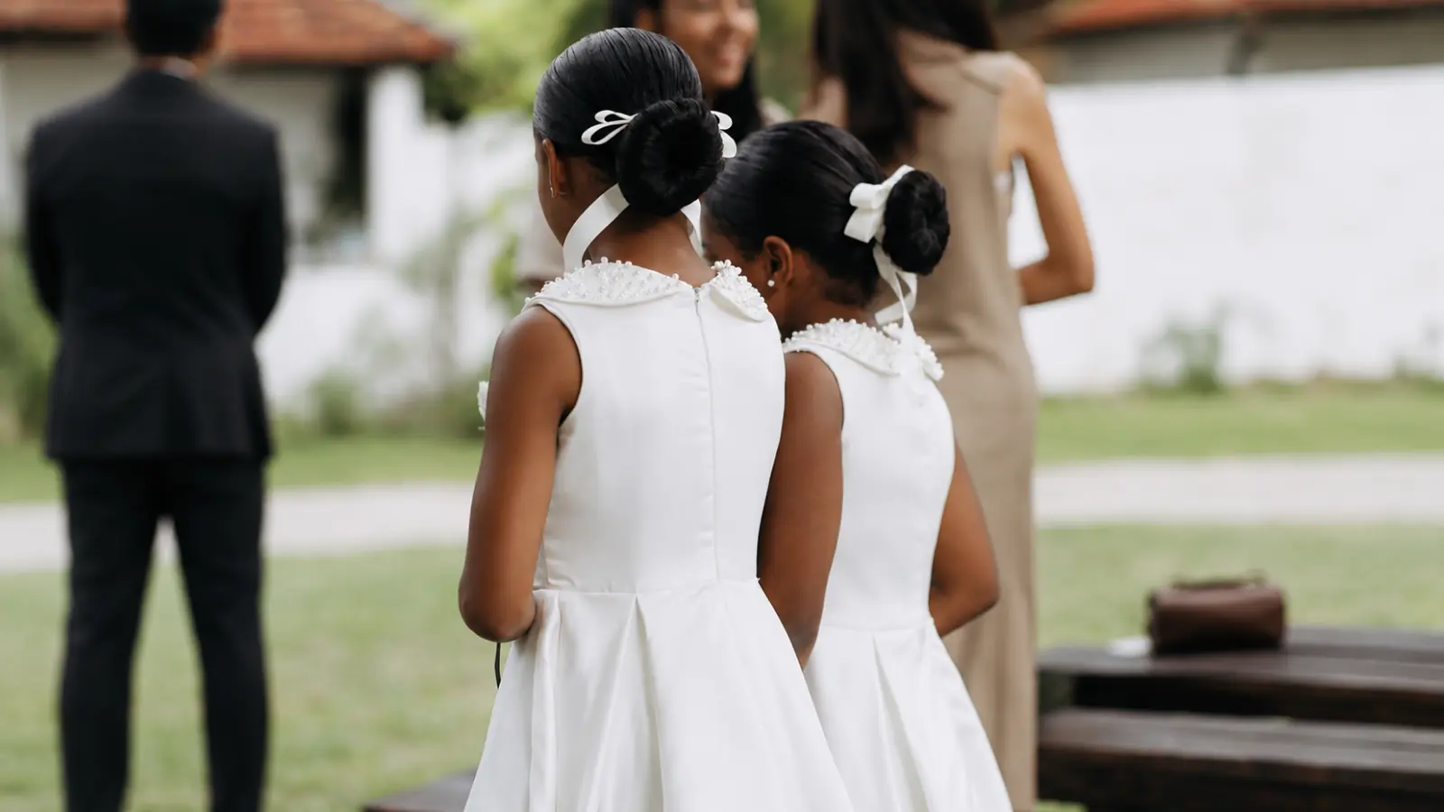 Two junior bridesmaids in coordinated white dresses with polished bun hairstyles standing together as younger attendants in an outdoor wedding party