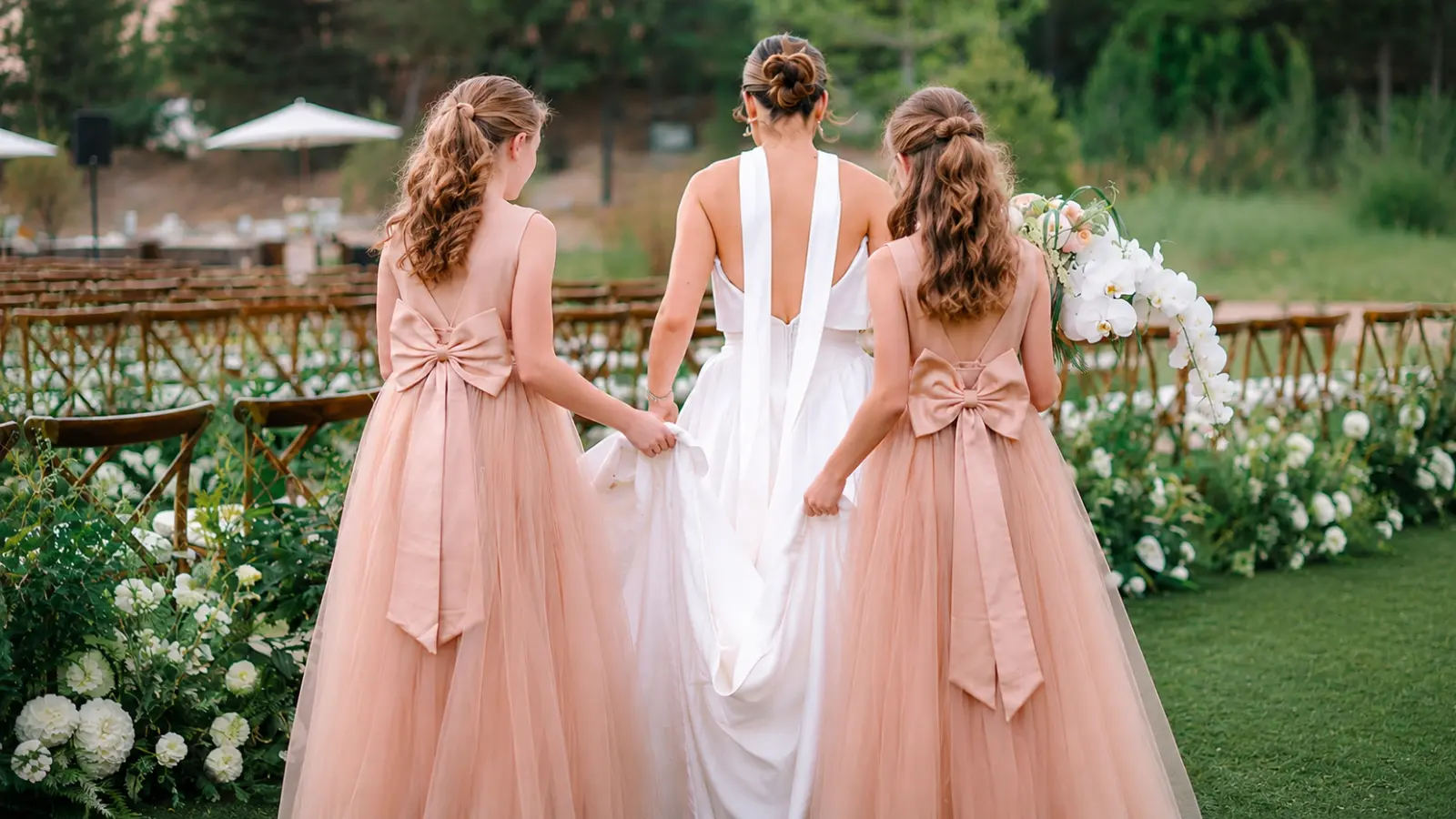 Junior bridesmaids wearing blush pink dresses and helping carry the bride's wedding gown while walking together toward an outdoor wedding ceremony