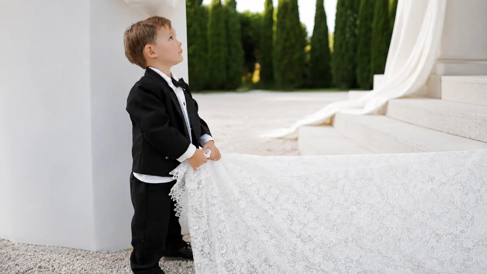 Little page boy in a formal black suit holding the bride’s long lace wedding dress train during an elegant outdoor wedding ceremony
