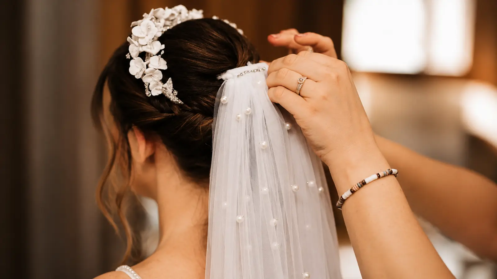 Pearl wedding veil being secured beneath a white floral bridal tiara on a dark hair updo during wedding preparation