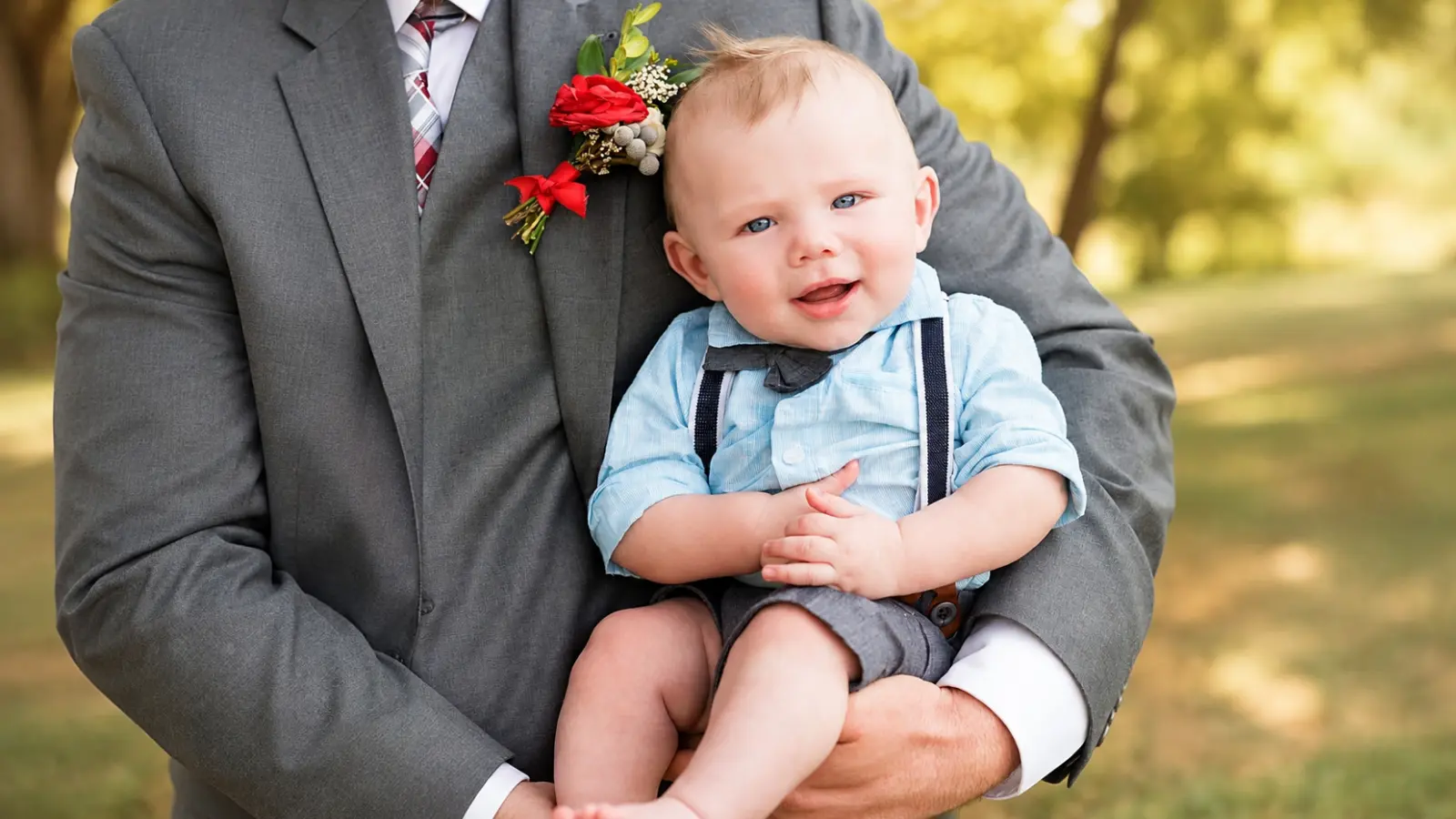 Baby ring bearer wearing suspenders and a bow tie is held by an adult, showing how infants can participate with help during a wedding Ring Bearer Age