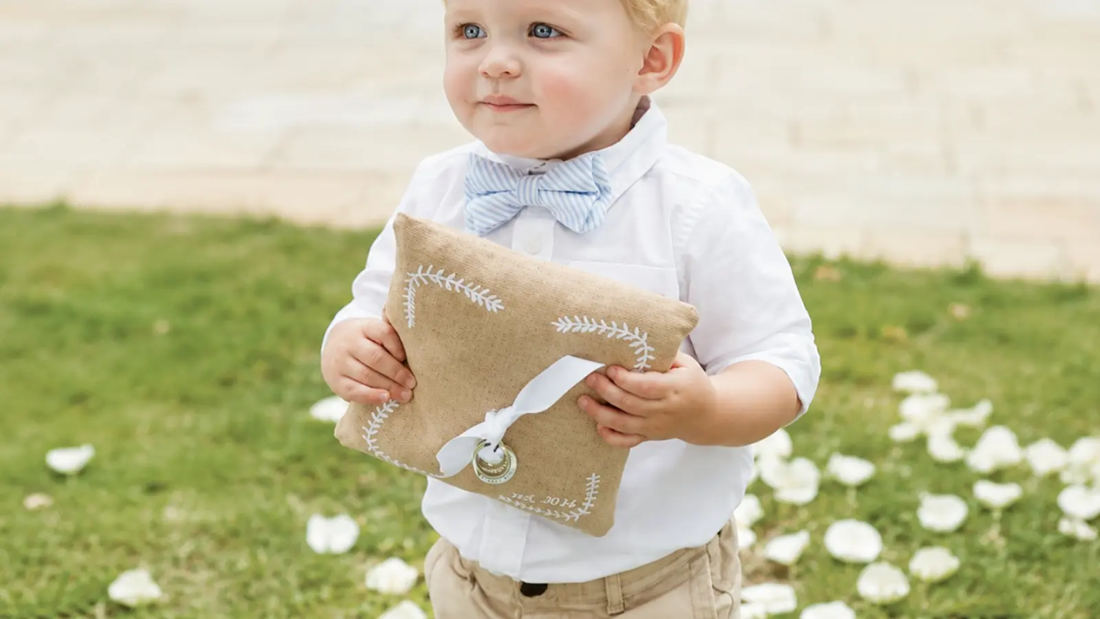 Toddler ring bearer wearing a bow tie carries a burlap ring pillow outdoors, showing how young children can participate in the wedding ceremony