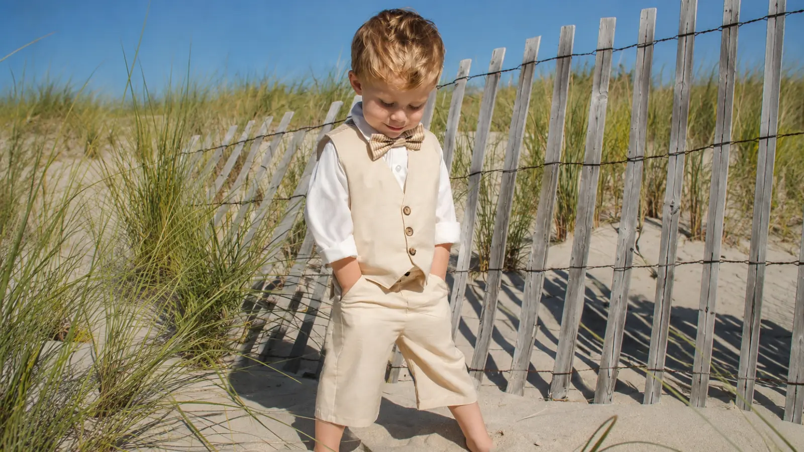 Young ring bearer wearing a beige vest and shorts with white shirt and striped bow tie for a casual beach wedding